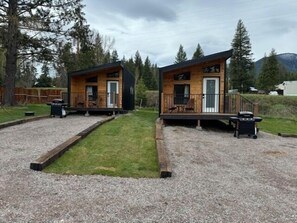 Dos cabañas de madera con terrazas separadas, cada una con una parrilla y mobiliario exterior.