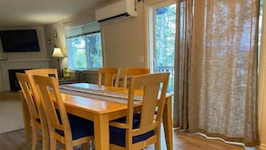 Een eethoek met een houten tafel en stoelen, een airconditioning, een open haard en uitzicht op bomen door het raam.