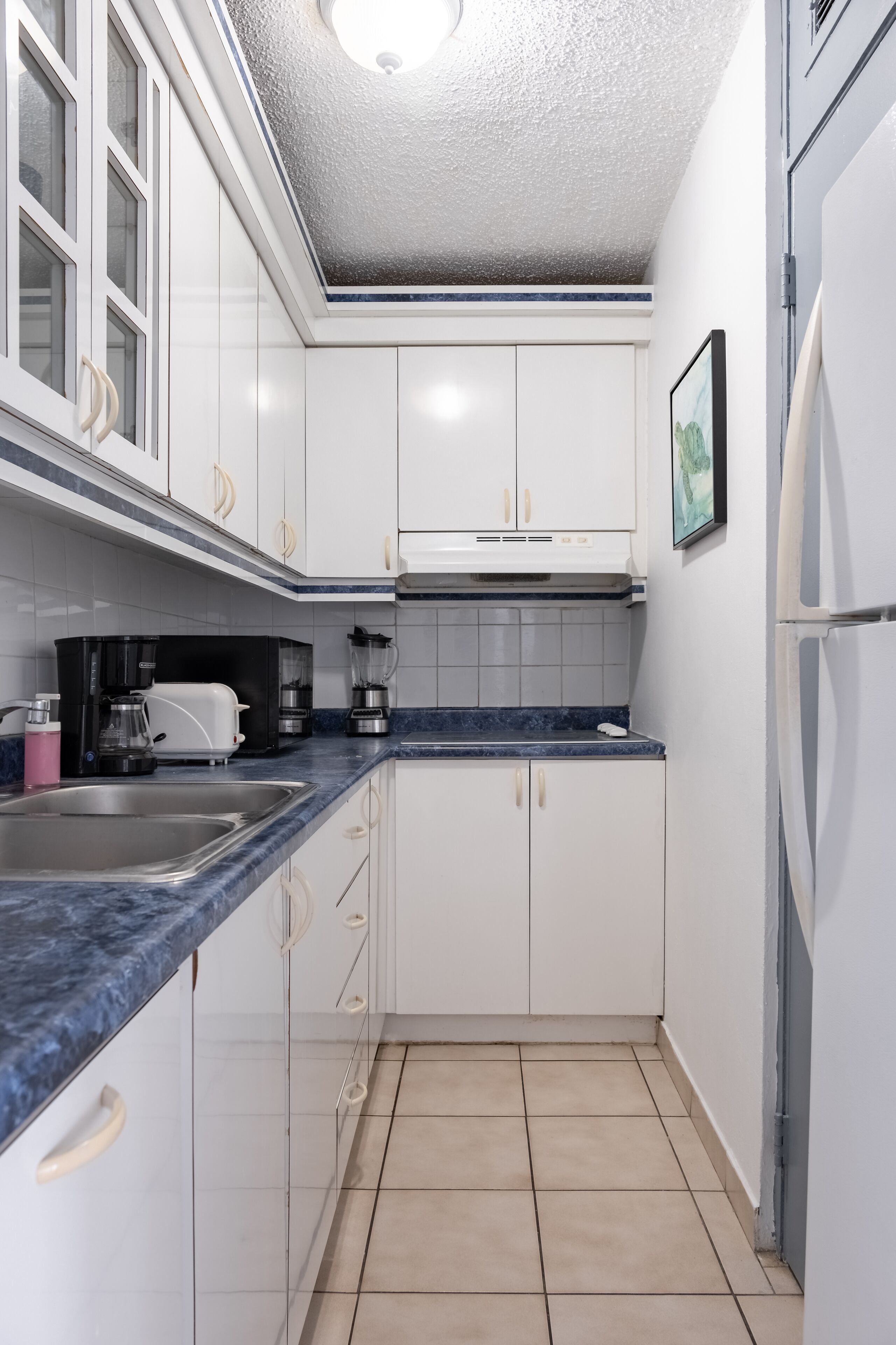 A compact kitchen with white cabinets, a blue countertop, a coffee maker, and a refrigerator.