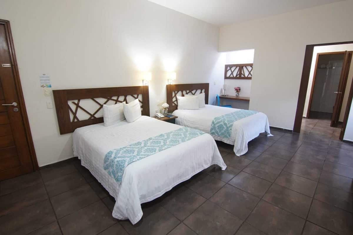 Habitación de hotel con dos camas, un cabecero de madera, una mesita de noche con lámpara y una pequeña mesa con un florero.