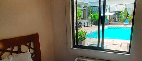 A bedroom with a large bed, a window with a view of a swimming pool and outdoor seating, and a built-in heater.