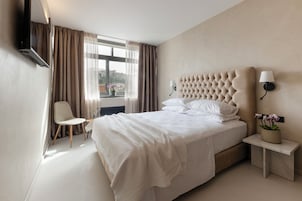 Quarto de hotel com uma cama grande, uma cadeira, uma mesa de cabeceira com abajur e uma TV fixada na parede.
