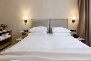 A hotel room with a large bed, two bedside lamps, a nightstand with a phone, and a shelf with folded towels and a vase of flowers.