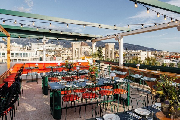 An outdoor dining area with tables and chairs, a view of the cityscape, and string lights overhead. - Regal Hotel Mitropoleos (Athens)