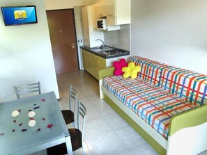 A compact living space with a kitchenette, a sofa with colorful cushions, a dining table with chairs, and a flat-screen TV mounted on the wall. - Fully Furnished Apartment on the top Floor (San Michele al Tagliamento)