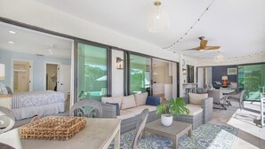 A spacious living area with a sofa set, a coffee table, and a dining table. There are large windows and a ceiling fan.