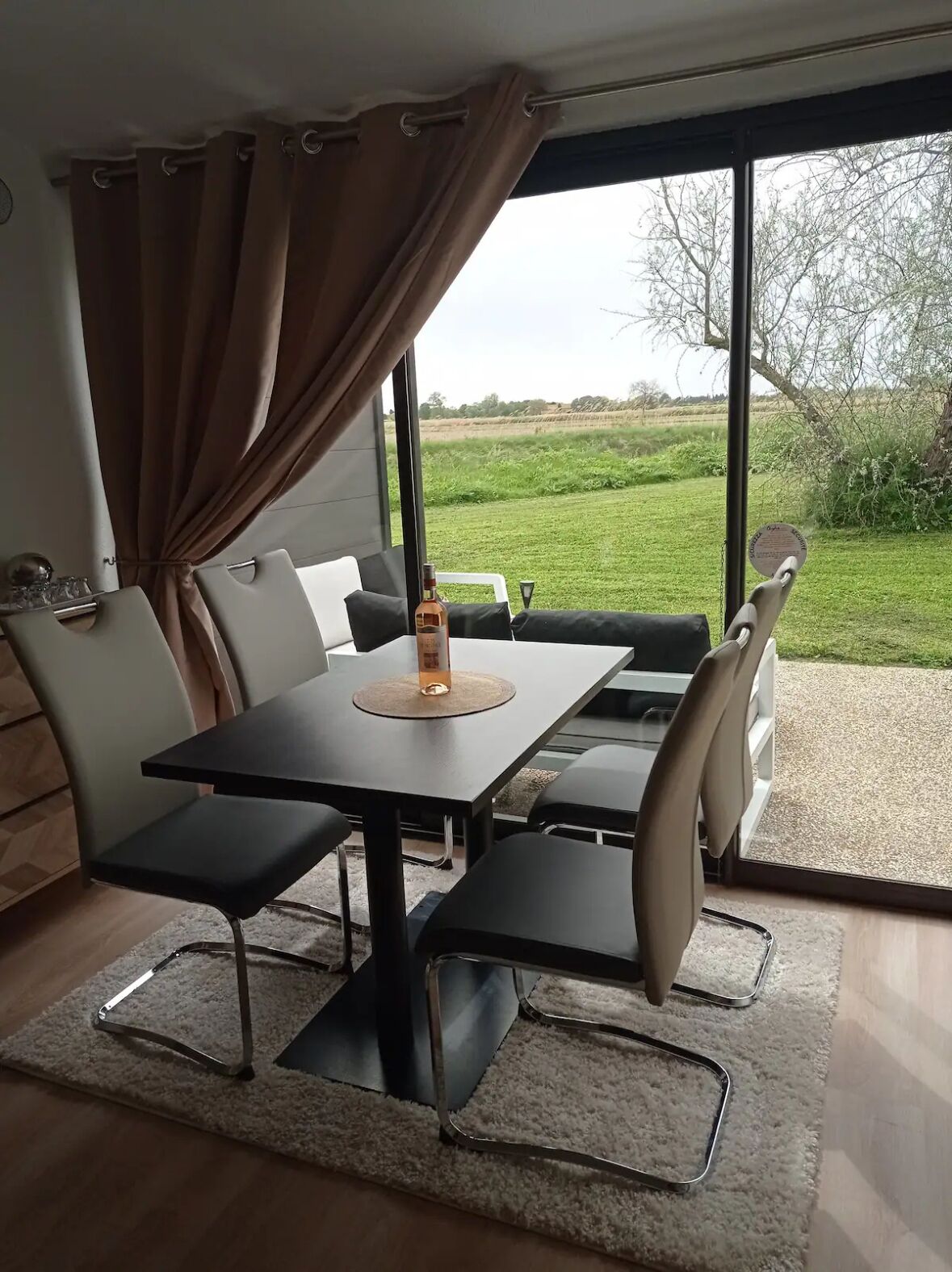 Uma área de refeições com mesa e cadeiras, uma garrafa de bebida alcoólica e vista para um campo gramado através de grandes janelas.