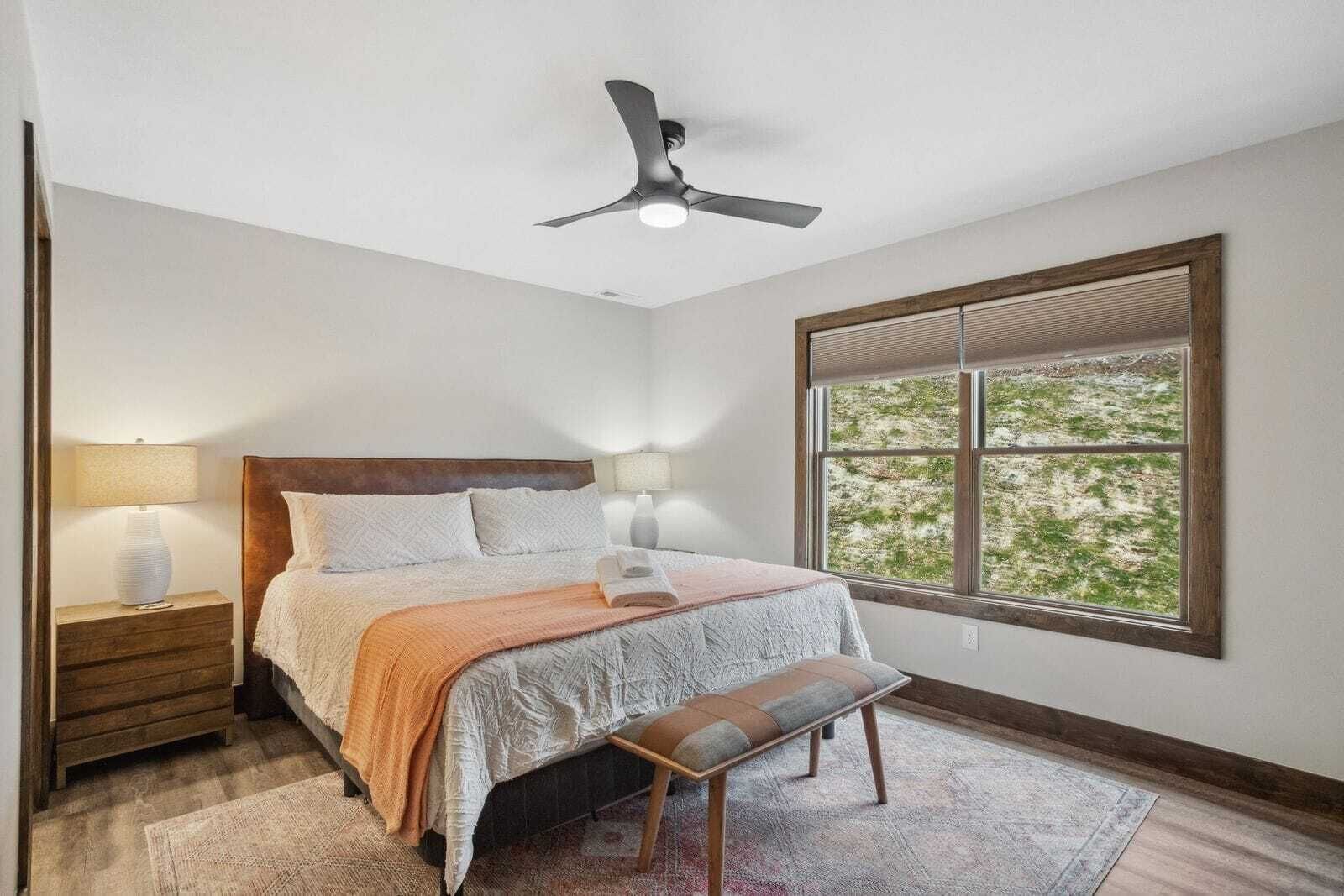 A bedroom with a bed, bedside lamps, a bench, a ceiling fan, and a window with a view of greenery.