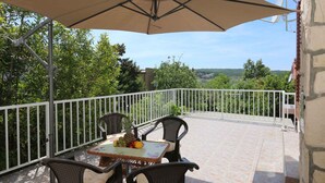 Varanda com mesa e cadeiras, um grande guarda-sol e vista para vegetação e colinas.