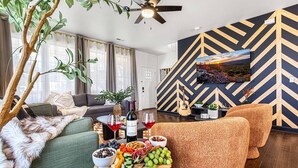 A modern living room with a fireplace, a dining area with a fruit and cheese board, and a large TV mounted on a chevron-patterned wall. - Mntn & Asheville Views! Movie Theater! Game Rm! Hot Tub! Fire Pit! Pets Welcome! (Candler)