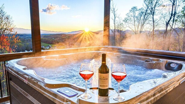 A hot tub with two glasses of red wine and a bottle, surrounded by a scenic view of trees and mountains. - Mntn & Asheville Views! Movie Theater! Game Rm! Hot Tub! Fire Pit! Pets Welcome! (Candler)