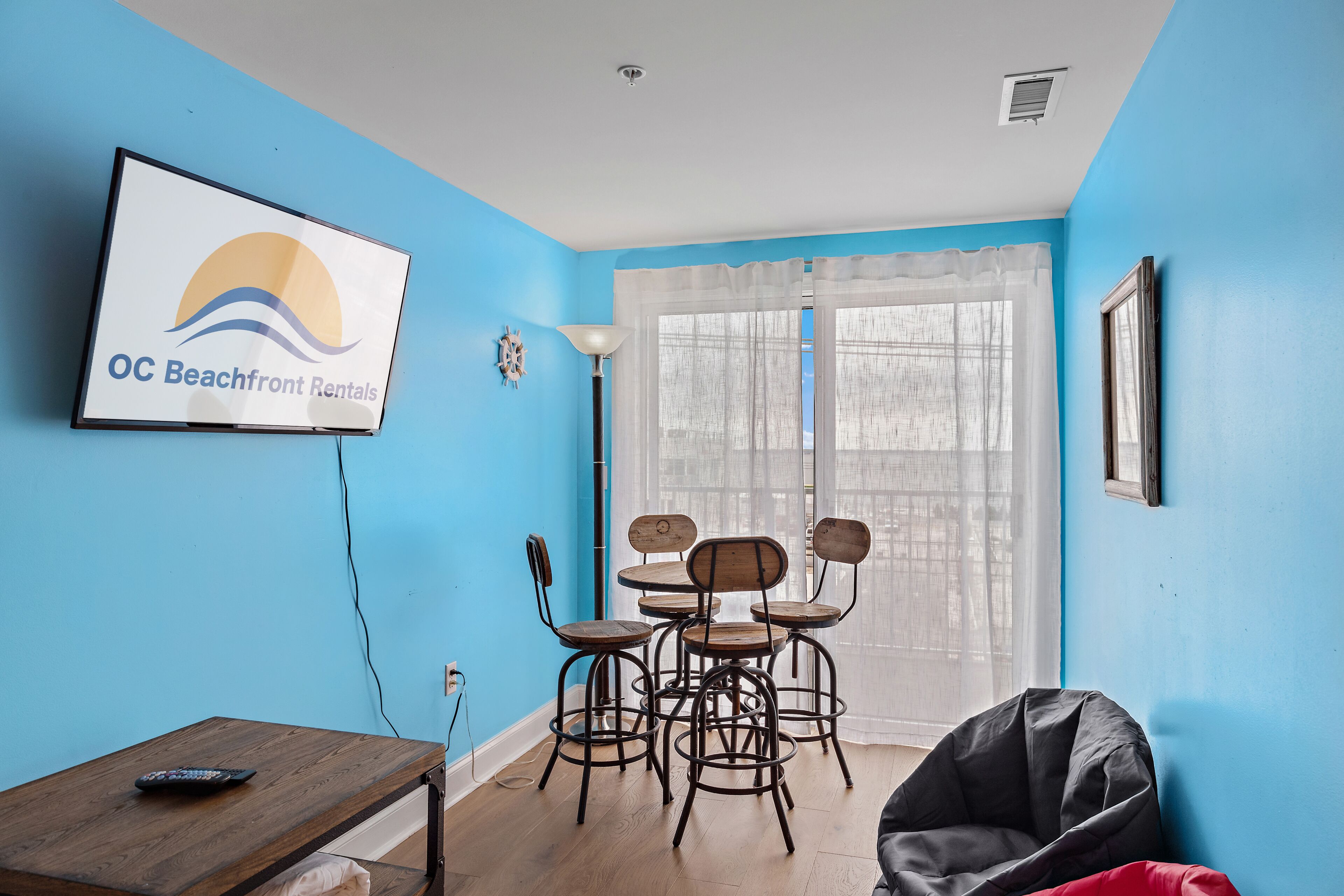 A room with a blue wall, a dining table with chairs, a TV mounted on the wall, and a window with sheer curtains.