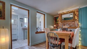 A dining area with a wooden table and chairs, a brick wall with a mirror, and a kitchen in the background. - Bulmer Cottage | Dog-Friendly Coastal Cottage, 10 Mins Walk to Beach (Winterton-on-Sea)