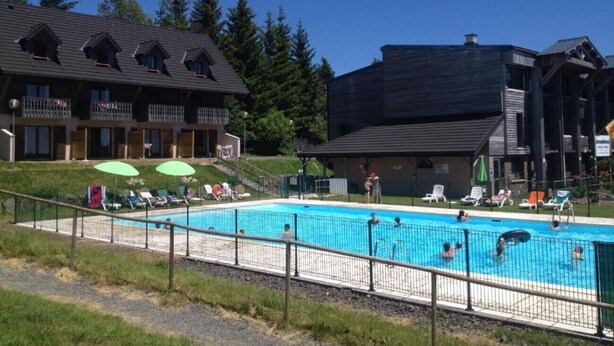 A swimming pool with lounge chairs and umbrellas, surrounded by buildings with balconies and a forested background.