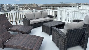 A rooftop terrace with wicker furniture, a white railing, and a view of buildings in the background.