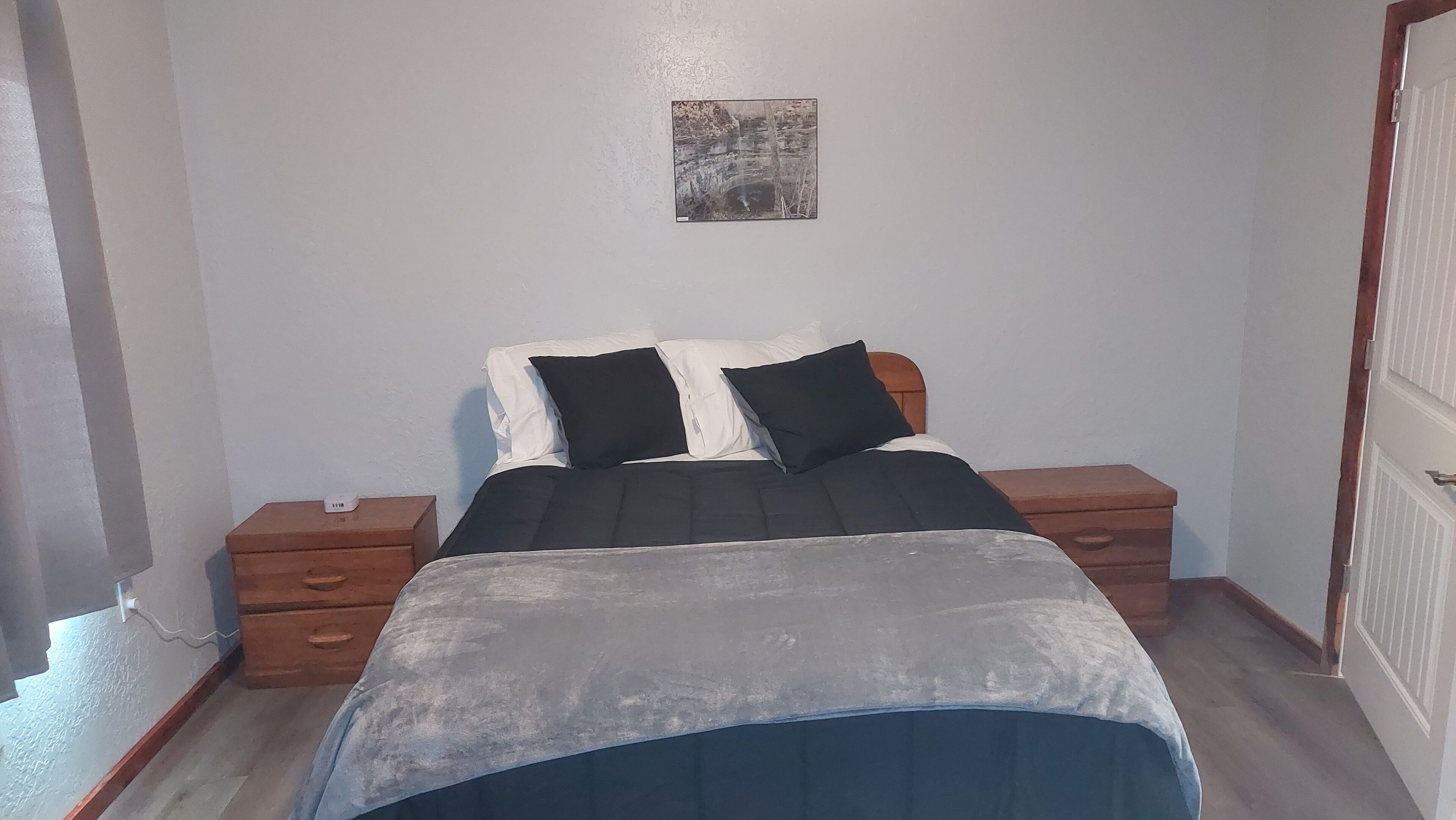 Una camera da letto con un letto, due comodini, un piccolo quadro appeso al muro e una porta.