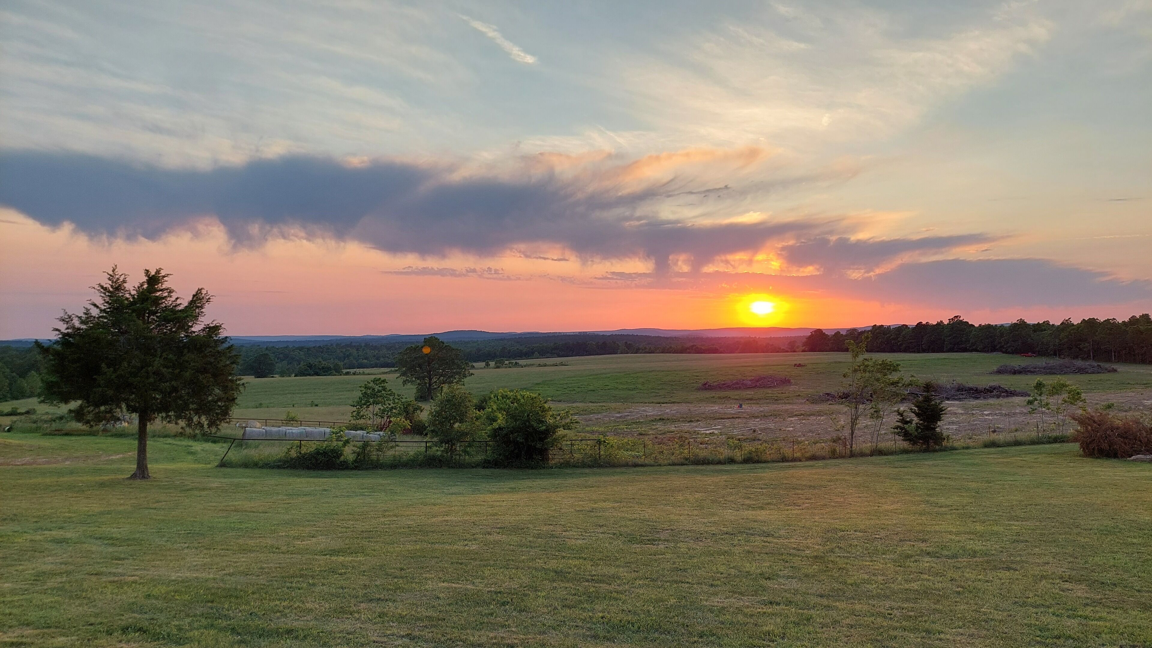 A sunset over a grassy field with a tree and hay bales.