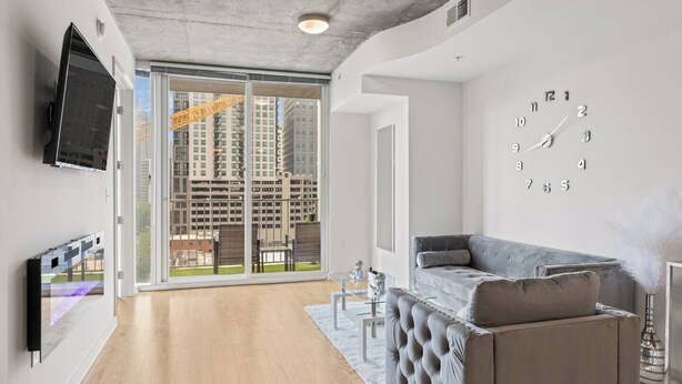 Un salón moderno con un sofá gris, un reloj de pared y un ventanal con vistas a la ciudad.