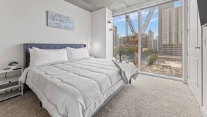 Un dormitorio con una cama grande, una mesita de noche, una planta en maceta, una lámpara de pared y vista a la ciudad a través de un ventanal corredera.