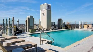 Una piscina en la azotea con sillones de descanso, un edificio alto al fondo y un cielo despejado.