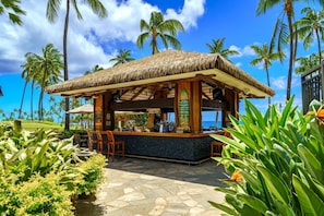 Un bar à toit de chaume, doté d’un sol en pierre, entouré de plantes tropicales et de palmiers.