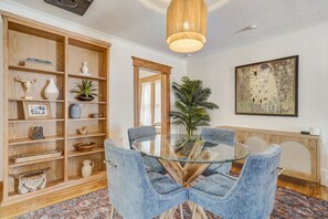Un coin repas avec une table en verre entourée de chaises bleues, une bibliothèque en bois contenant divers objets, un grand tableau accroché au mur et un luminaire suspendu.