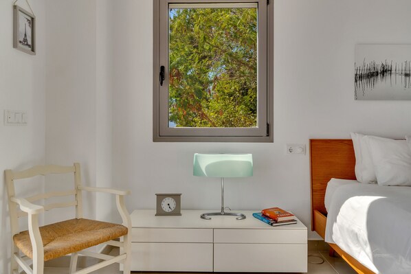 A bedroom with a bed, a bedside table with a lamp, a chair, and a window with a view of trees.