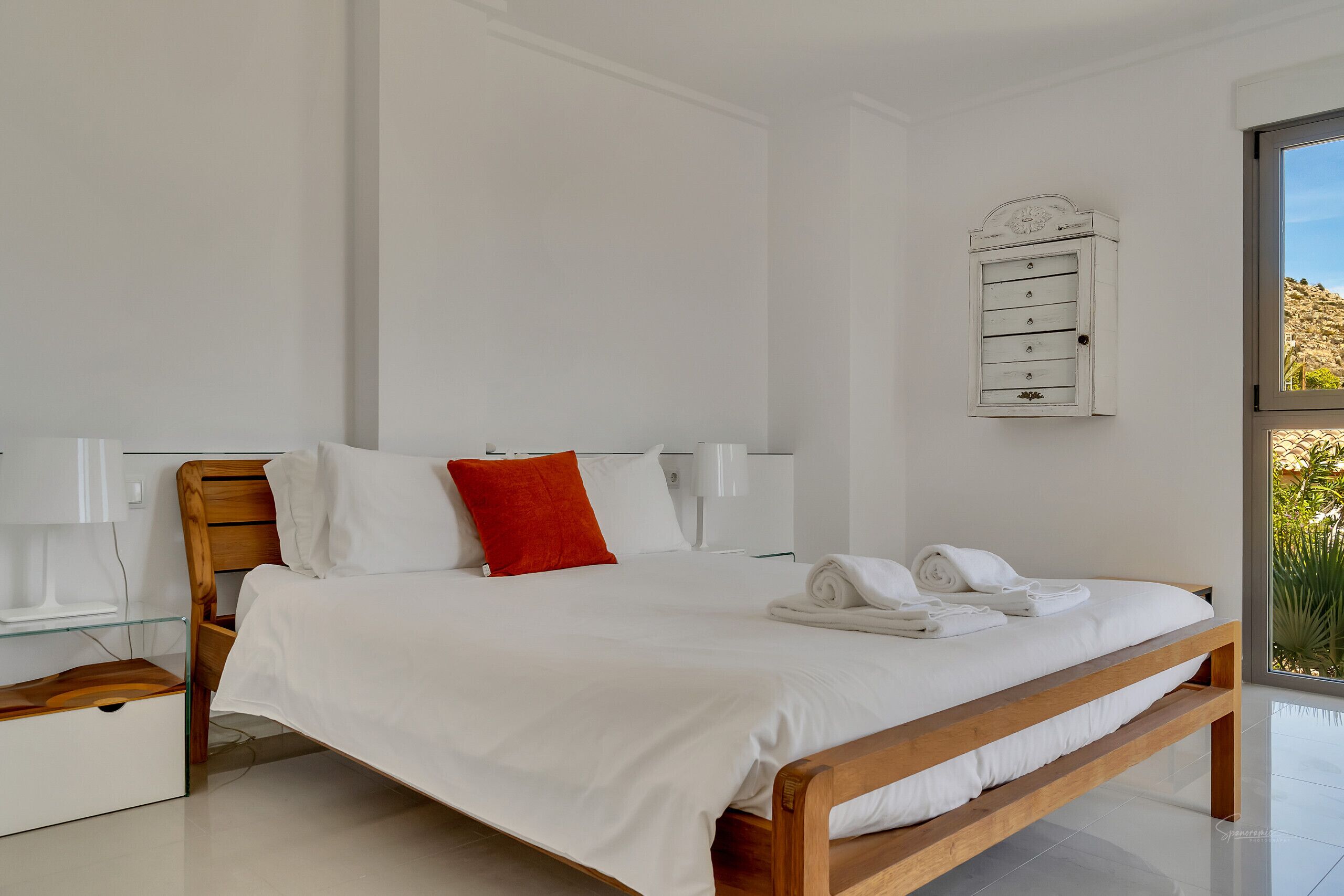 A modern bedroom with a wooden bed, white bedding, a red pillow, and a white bedside table.