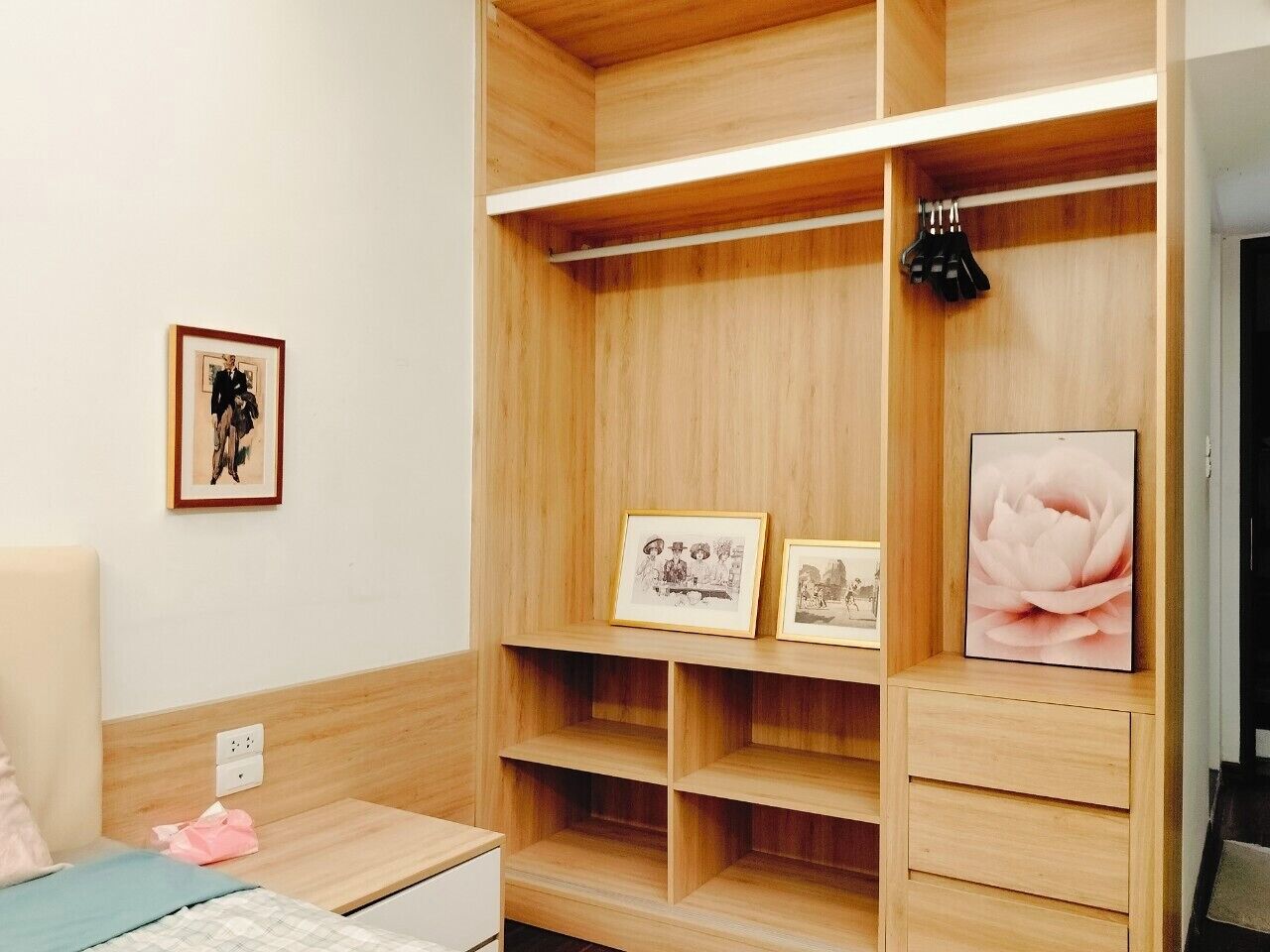A neatly organized room with a wooden wardrobe, a bed, and a desk. There are framed pictures on the wall and a pink flower painting on the wardrobe.
