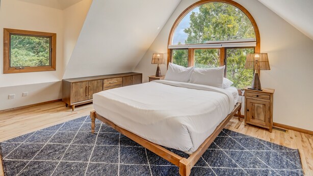 Un dormitorio con una cama grande, cabecera de madera, dos mesitas de noche con lámparas, una ventana con vistas a árboles y suelo de madera.