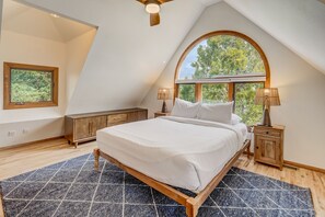 A bedroom with a large bed, wooden headboard, two bedside tables with lamps, a window with a view of trees, and a wooden floor.