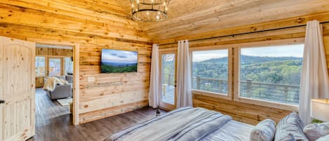A wooden cabin bedroom with a large bed, a television, and a scenic view through the windows.