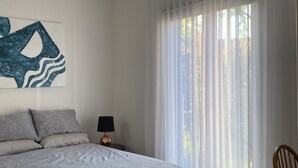 Un dormitorio con cama, mesita de noche, lámpara, silla y un amplio ventanal con cortinas traslúcidas.