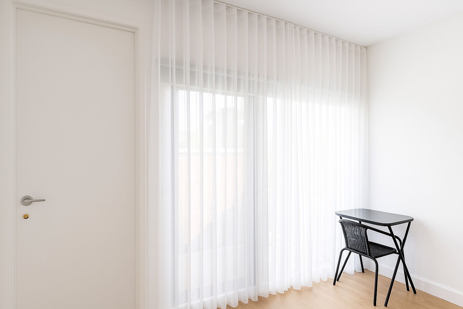 A room with a white door, sheer white curtains, a small black table with a chair, and a wooden floor.