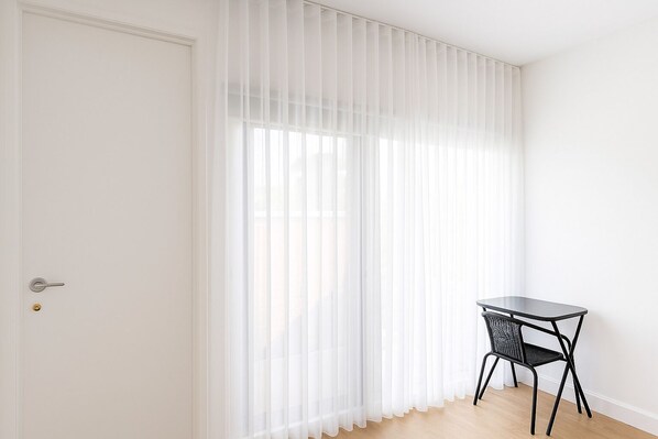 A room with a white door, sheer white curtains, a small black table with a chair, and a wooden floor.