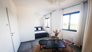 Une pièce de taille modeste comprenant un lit, une petite table avec des chaises, un vase de fleurs et une vue sur l’extérieur par une fenêtre.