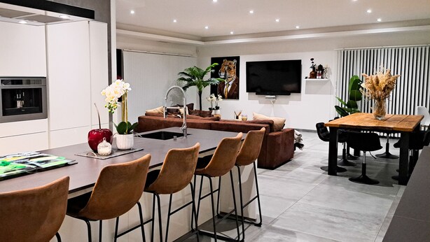Une cuisine moderne dotée de fours encastrés, un îlot central avec des tabourets de bar, un coin repas avec une table et des chaises en bois, et un espace de vie avec un canapé et un téléviseur fixé au mur.