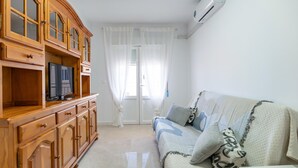 Ein Wohnzimmer mit einem Holzschrank, einem Fernseher, einer Couch mit Kissen und einem Fenster mit durchsichtigen Vorhängen.
