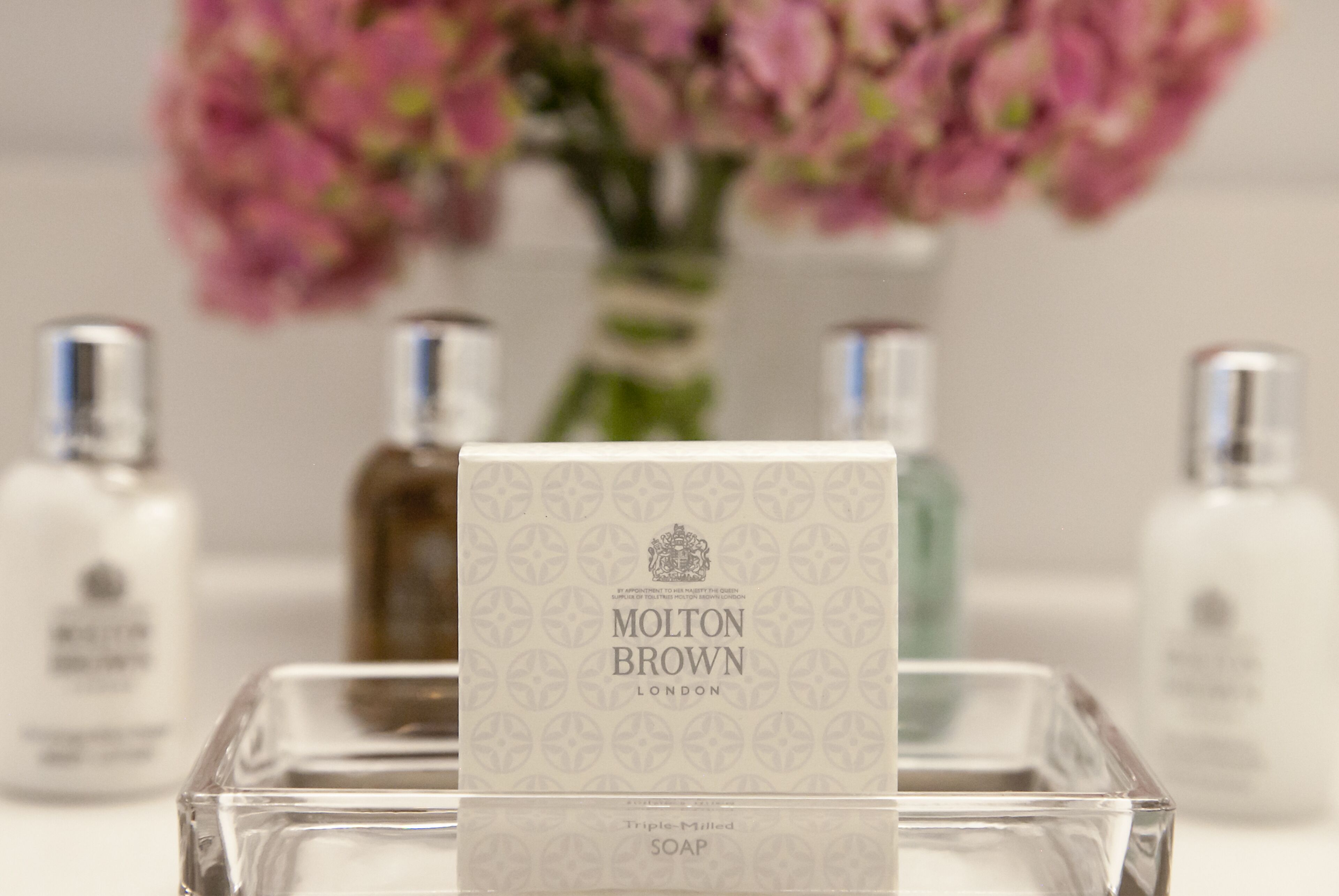 En klar glasflaske med sæbe, mærket 'Molton Brown London', foran en sløret baggrund af blomster og andre flasker.