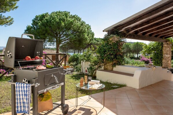 An outdoor grill with food, a table with wine and bread, and a view of the sea.