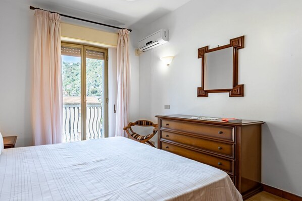 A bedroom with a bed, a dresser, a chair, a window with curtains, and an air conditioning unit.
