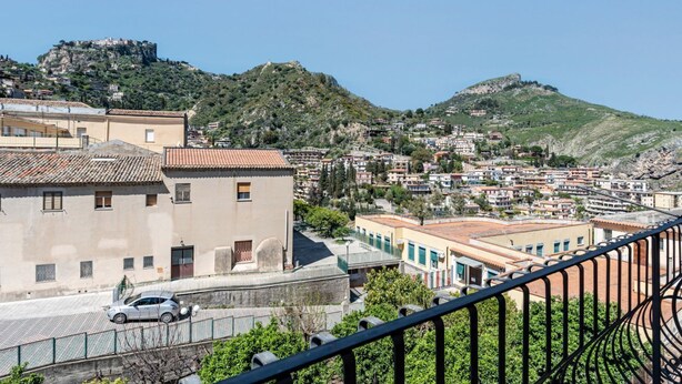A coastal town with buildings, a prominent hill with a fort, and a clear sky. - GuestHost - Spacious 81 sqm apartment in Taormina, ideal for 6 guests. Featuring two terraces from which to enjoy breathtaking sea views, this accommodation offers a unique experience. Perfectly situated just a short walk from the vibrant center of Taorm (Taormina)