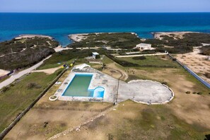 An aerial view of a property with a swimming pool, a tennis court, and a large open area with a view of the ocean.