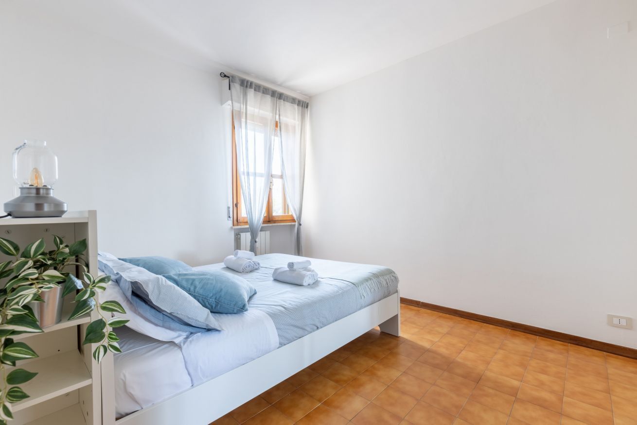 A bedroom with a bed, a bedside table with a lamp, a window with sheer curtains, and a potted plant.