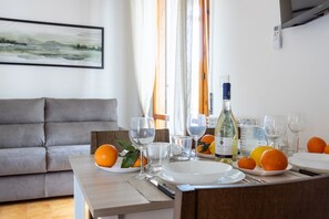 A dining table set with plates, glasses, and a bottle of wine, with a grey sofa and a painting in the background.