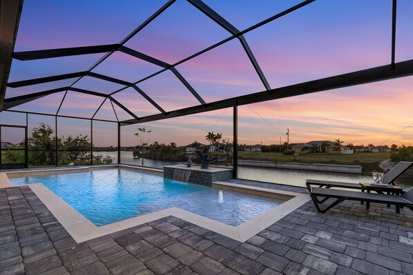 An outdoor swimming pool with a view of the sunset, surrounded by lounge chairs and a waterfall feature. - Gulf Access, Heated Saltwater Pool, Kayaks - Villa Sunset Lagoon (Cape Coral)