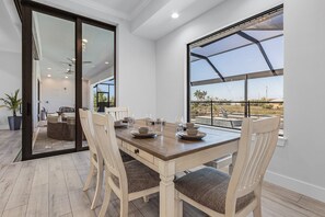 A dining area with a wooden table, four chairs, and a large window with a view of the outdoors. - Gulf Access, Heated Saltwater Pool, Kayaks - Villa Sunset Lagoon (Cape Coral)