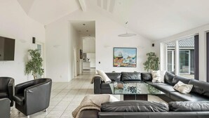 A modern living room with a black leather sofa, a glass coffee table, and a TV mounted on the wall. There are two potted plants and a painting on the wall. - Beautiful home in Jerup with sauna (Jerup)