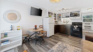 Een kleine keuken met witte kasten, een inbouwmagnetron en een zwart fornuis. Er staat een rond tafel met twee stoelen en een klein plankje met boeken.
