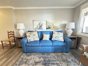 Ein blaues Sofa mit gemusterten Kissen, zwei Beistelltische mit Lampen, ein Holzstuhl und ein Teppich.